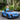Child in a blue Audi toy car outdoors with greenery in the background