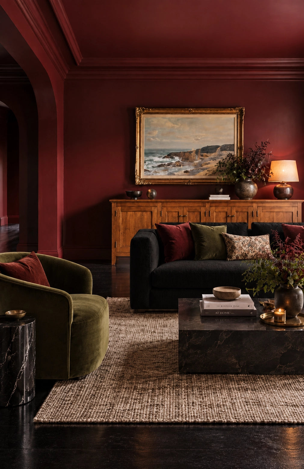 Burgundy living room with black fabric sofa, black marble coffee and side tables, olive green lounge chair, wood credenza, neutral rug, and gold-framed coastal artwork.