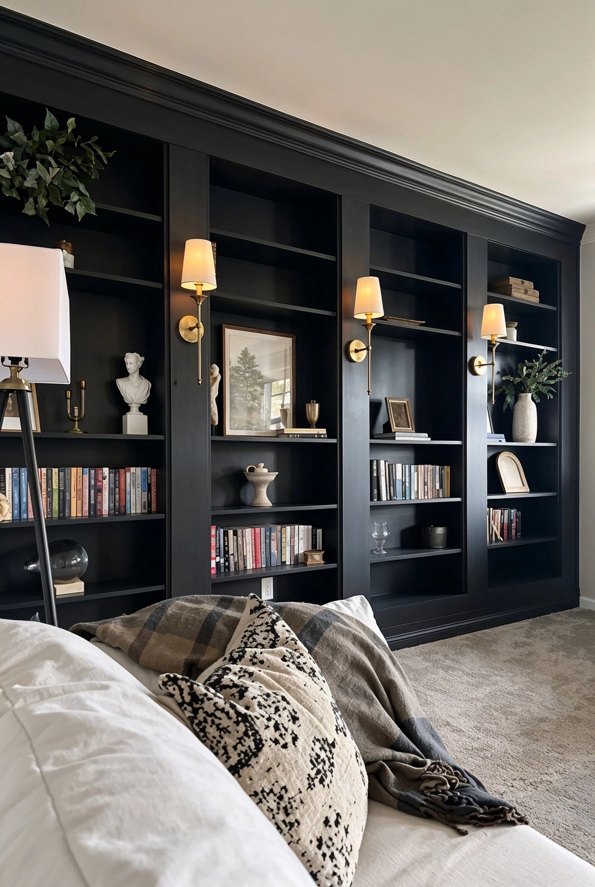 Black living room ideas with matte black built-in shelving, brass wall sconces, books and artwork, cream sofa, patterned neutral pillows, and pale area rug.
