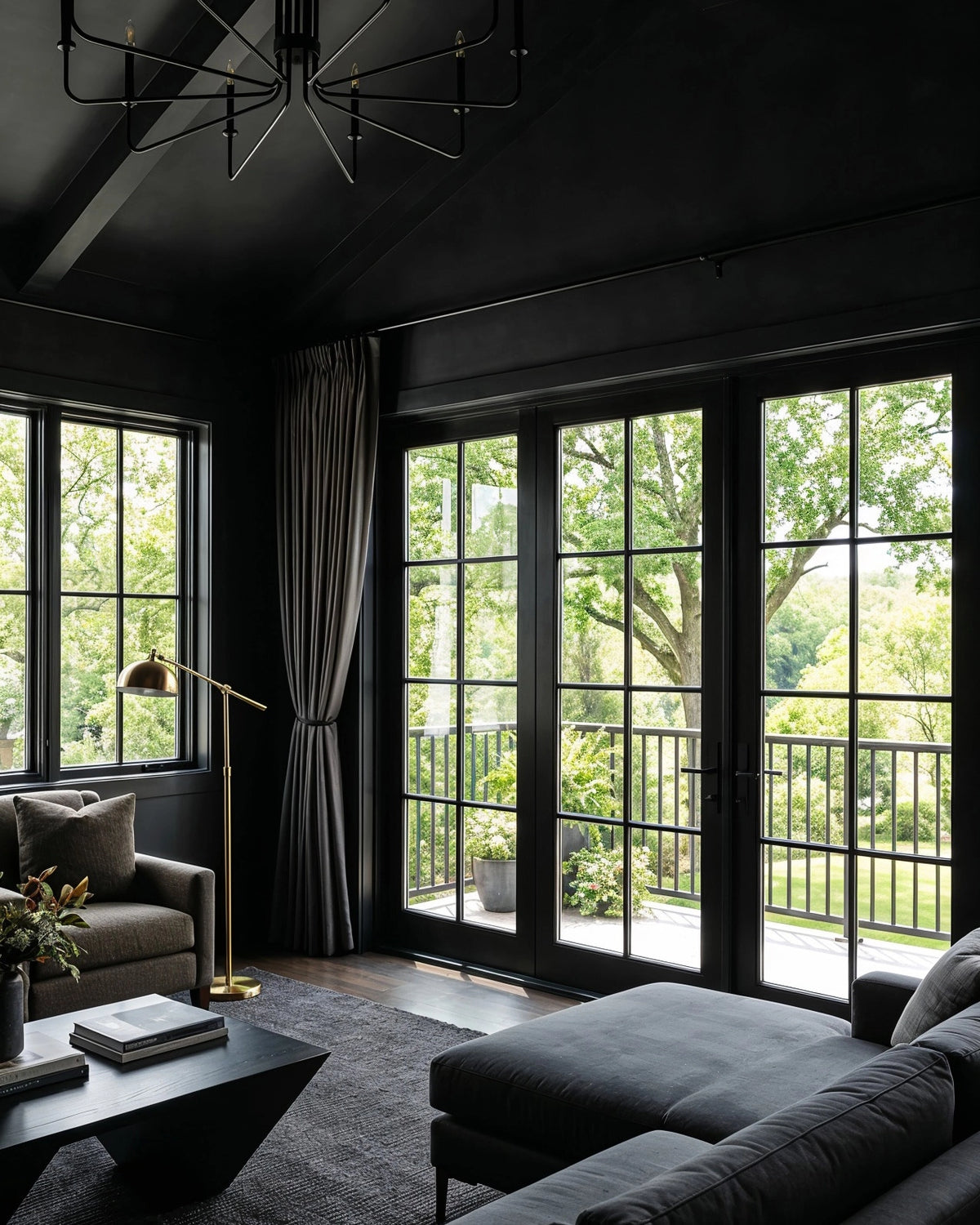 Black living room ideas with black-framed French doors and windows, vaulted dark ceiling, charcoal sectional sofa, black coffee table, brass floor lamp, and green outdoor views.