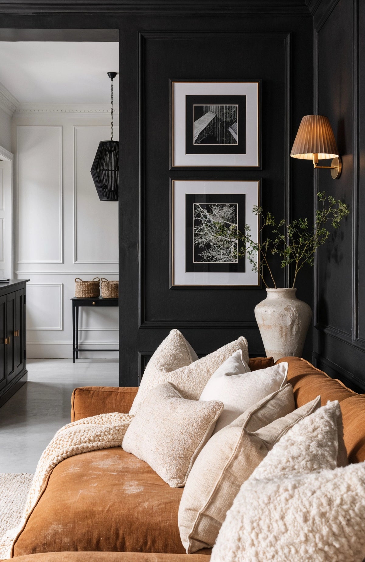 Black living room ideas with paneled black walls, cognac sofa, cream boucle and linen pillows, framed black-and-white art, and a warm wall sconce.