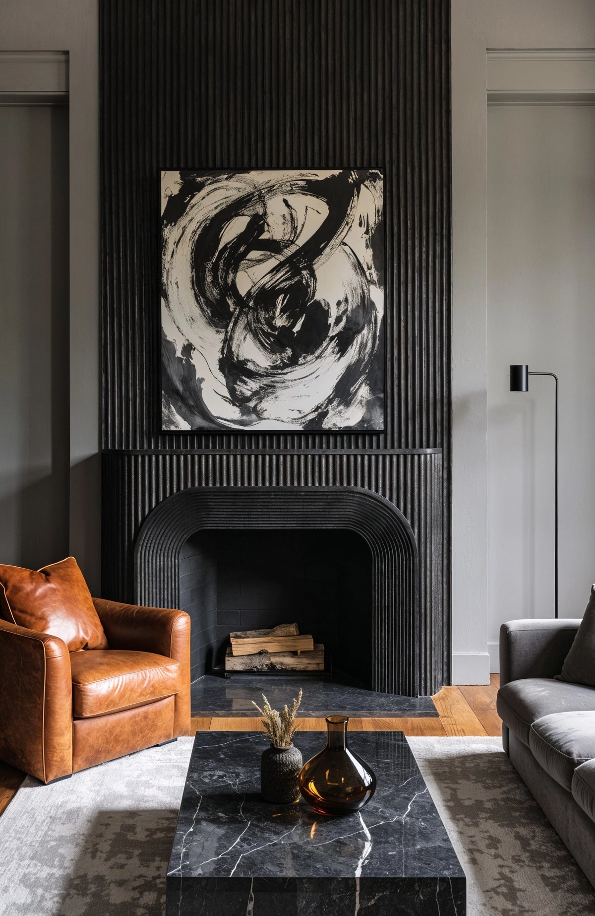 Black living room ideas with a sculptural ribbed black fireplace, black and white abstract artwork, cognac leather chair, gray sofa, and black marble coffee table.