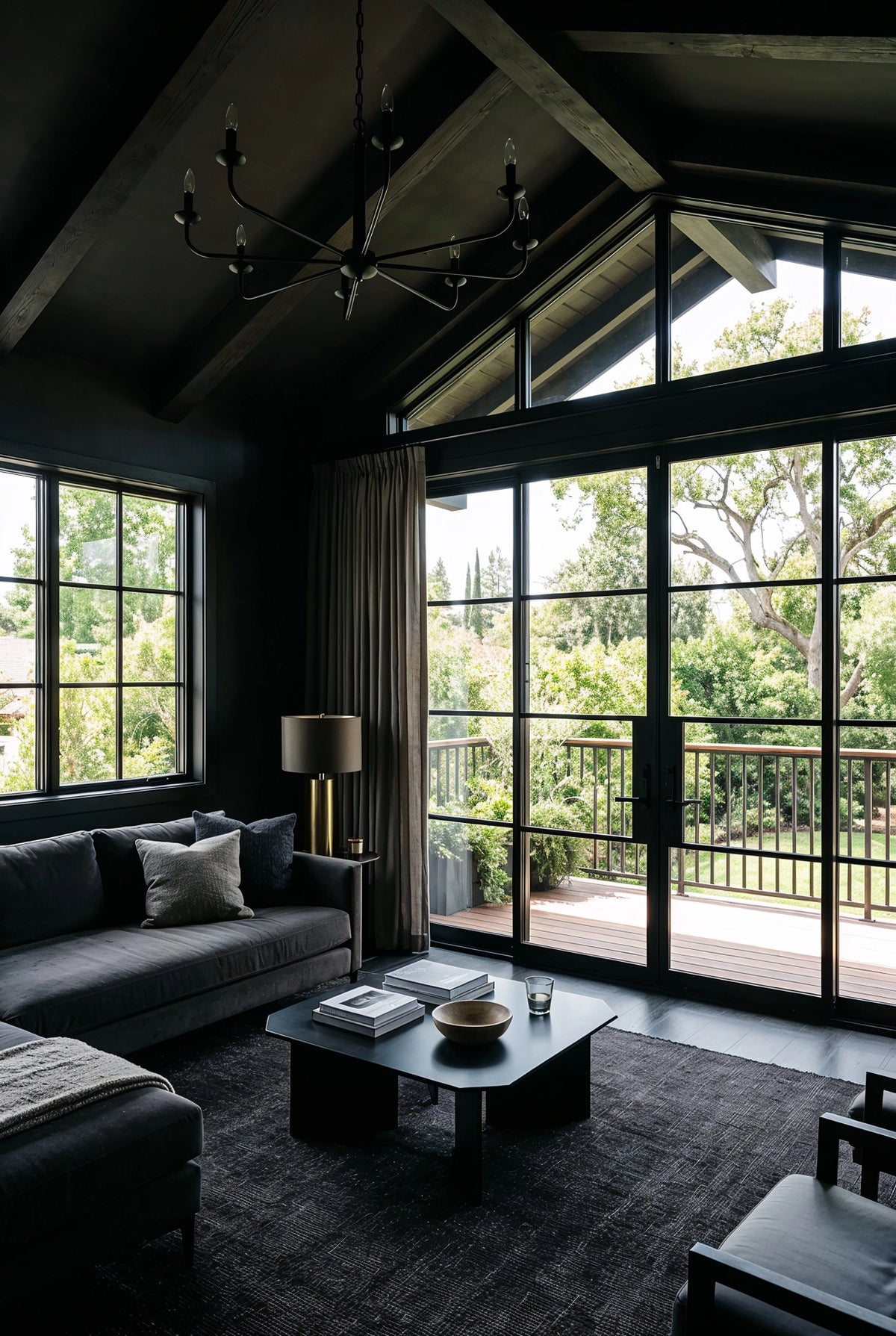 Black living room ideas with vaulted ceiling, exposed dark beams, floor-to-ceiling black-framed windows and doors, charcoal sofa, black coffee table, brass floor lamp, and garden views.