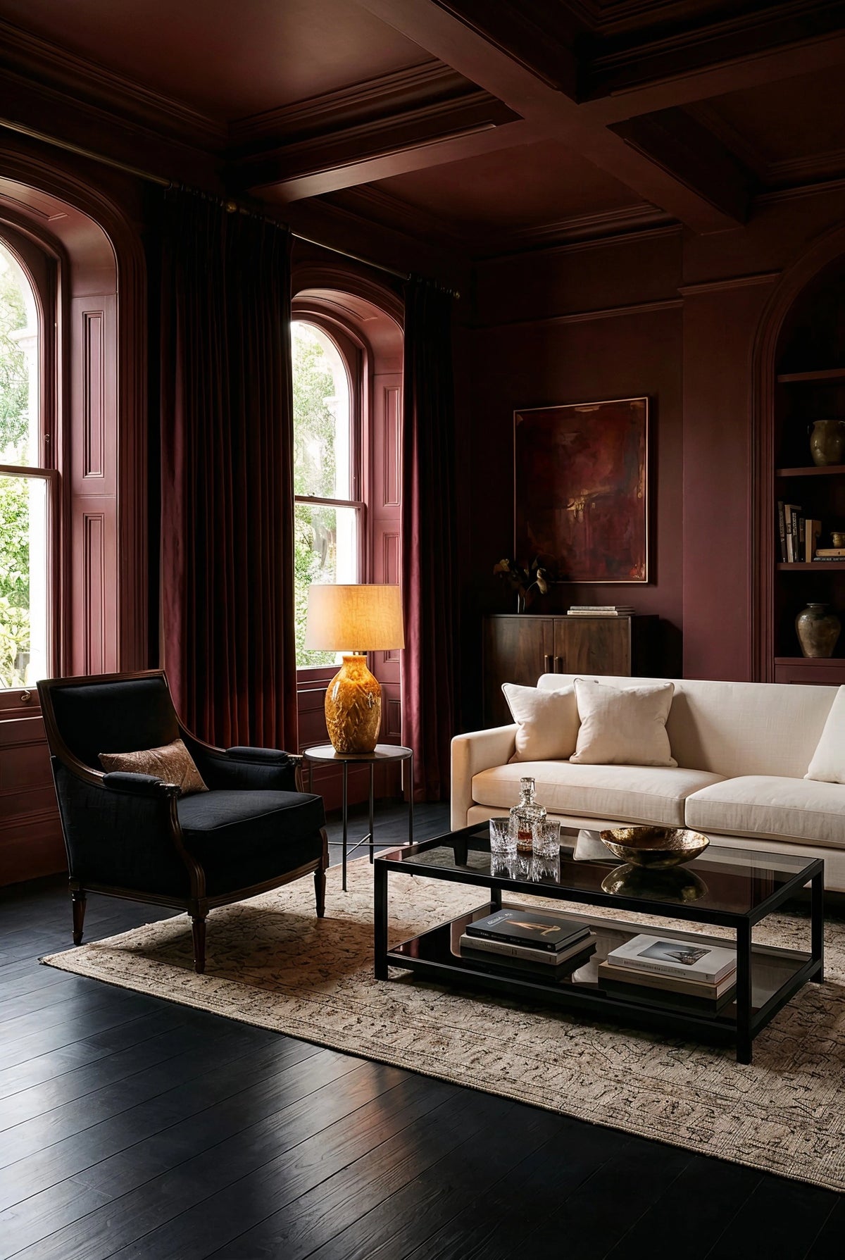 Oxblood living room with black wood floor, black lounge chair, cream sofa, arched windows, dark curtains, black coffee table, and warm amber lighting.