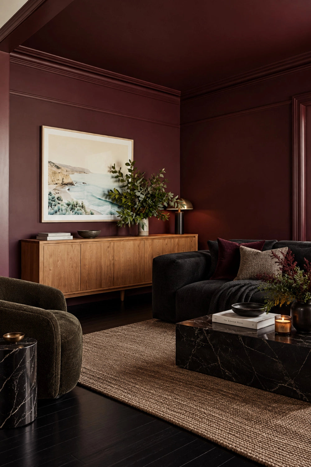 Oxblood living room with black fabric sofa, black marble coffee table, dark olive lounge chair, natural wood credenza, woven neutral rug, and coastal wall art.