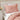 Pink textured pillow and blanket on a bed with a nightstand and flowers in the background.