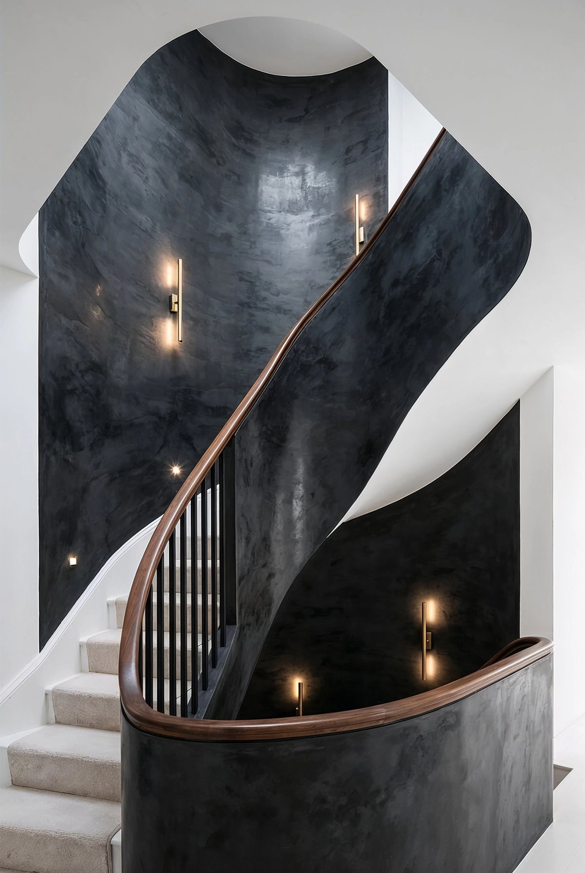 Curved staircase with textured black plaster walls, warm wood handrail, black balusters, brass wall sconces, white architecture, and pale carpeted steps.